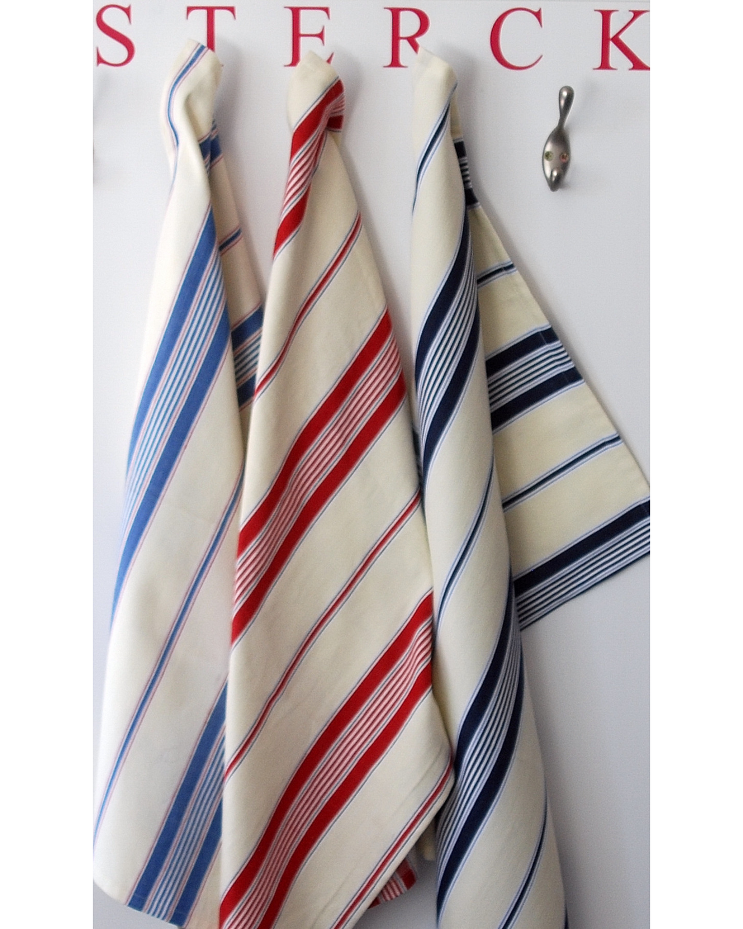 three striped cotton tea towels hanging on hooks. the tea towels are made from pure cotton and are a beige fabric with blue, navy and red stripes.