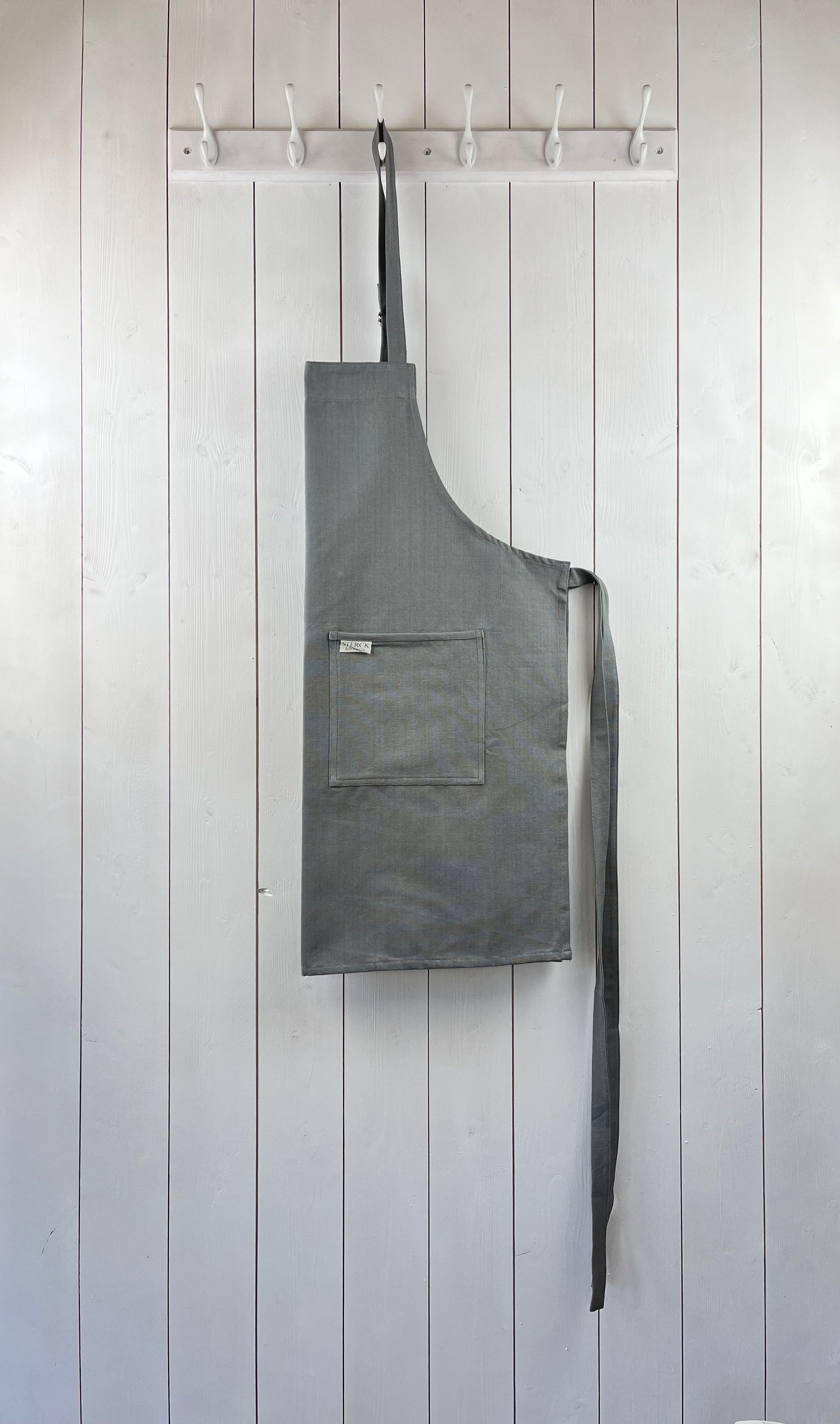 A grey cotton apron with double front pockets and adjustable neck strap. Sterck & Co.