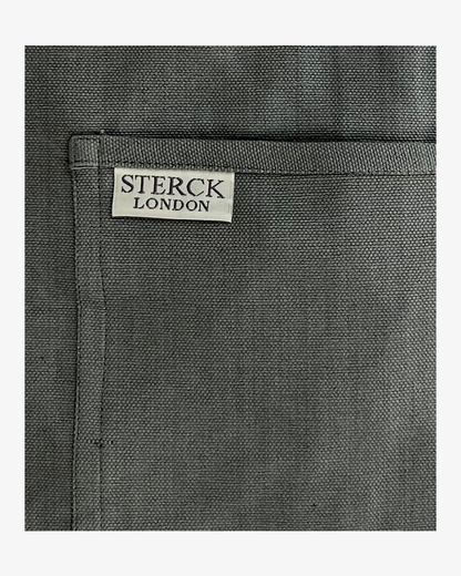 A grey cotton apron with double front pockets and adjustable neck strap. Sterck & Co. Close up of fabric and pocket detailing.