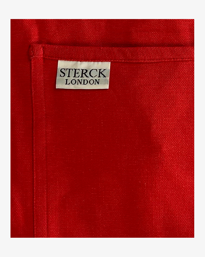 a poppy red cotton apron with double front pockets and adjustable neck strap. sterck & co. close up of fabric and pocket detailing.