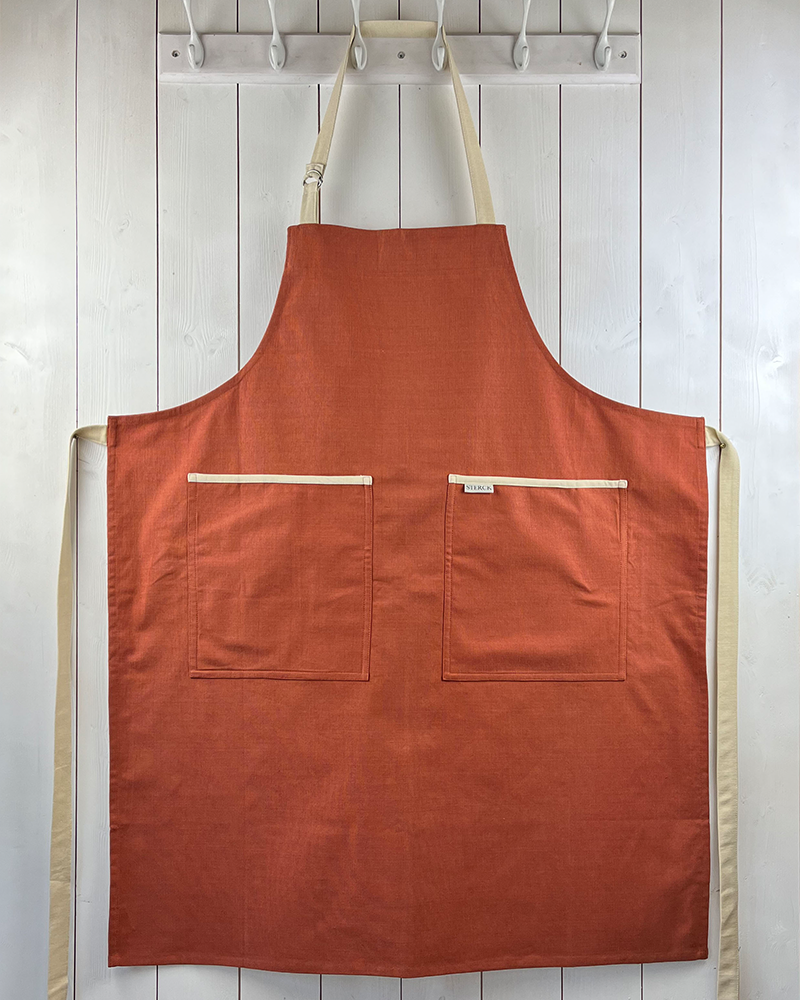 A dusky red apron with cream detailing, double front pockets and adjustable neck strap. Sterck & Co.