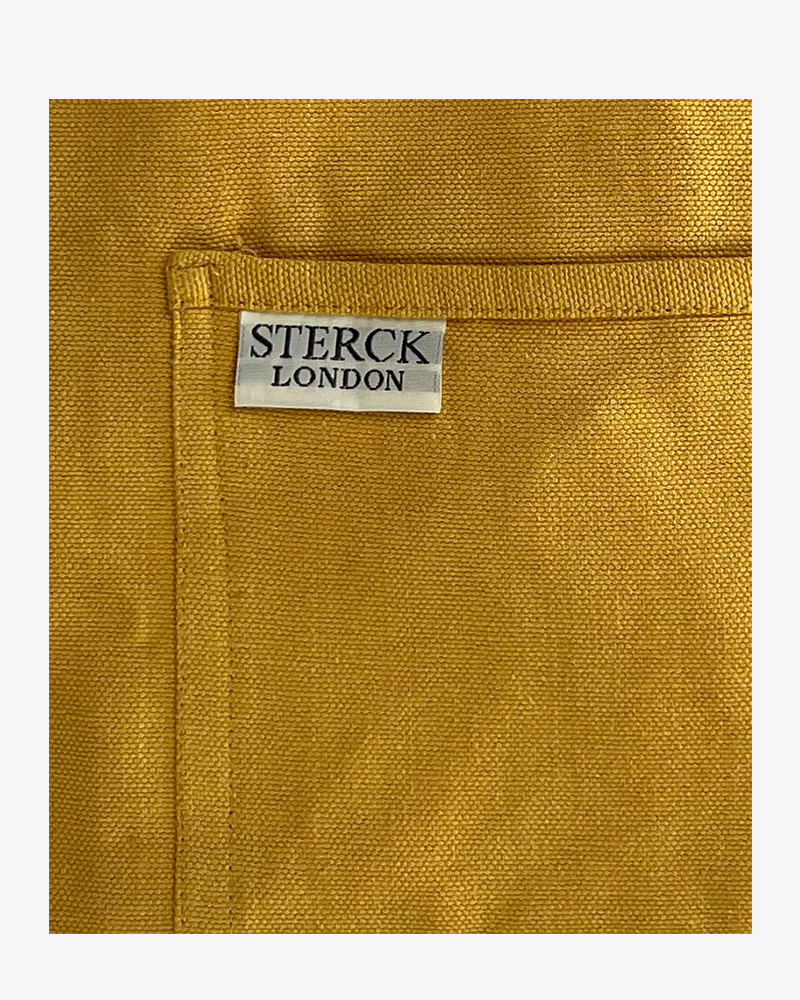 A sunflower yellow cotton apron with double front pockets and adjustable neck strap. Sterck & Co. Close up of fabric and pocket detailing.