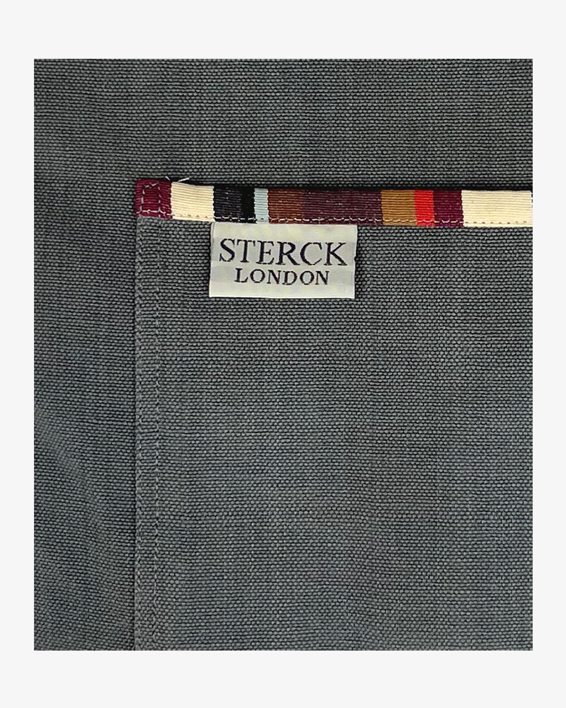 A bold grey cotton apron enlivened by stripey detailing. Double front pockets, stripy ties and adjustable neck strap. Sterck & Co. Close up of fabric and pocket detailing.