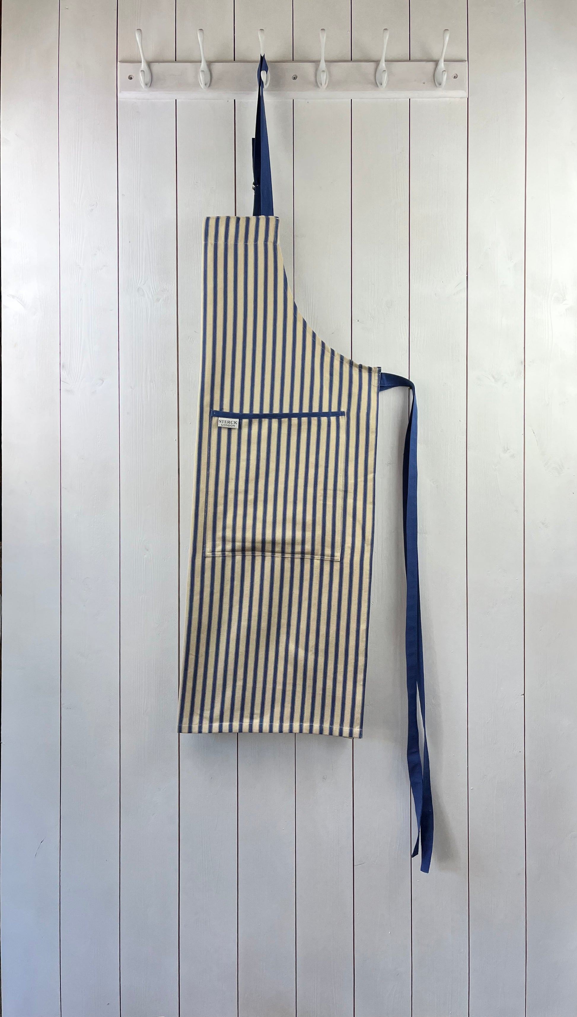 A classic cream and blue striped cotton apron with double front pockets, blue ties and adjustable neck straps. Sterck & Co.