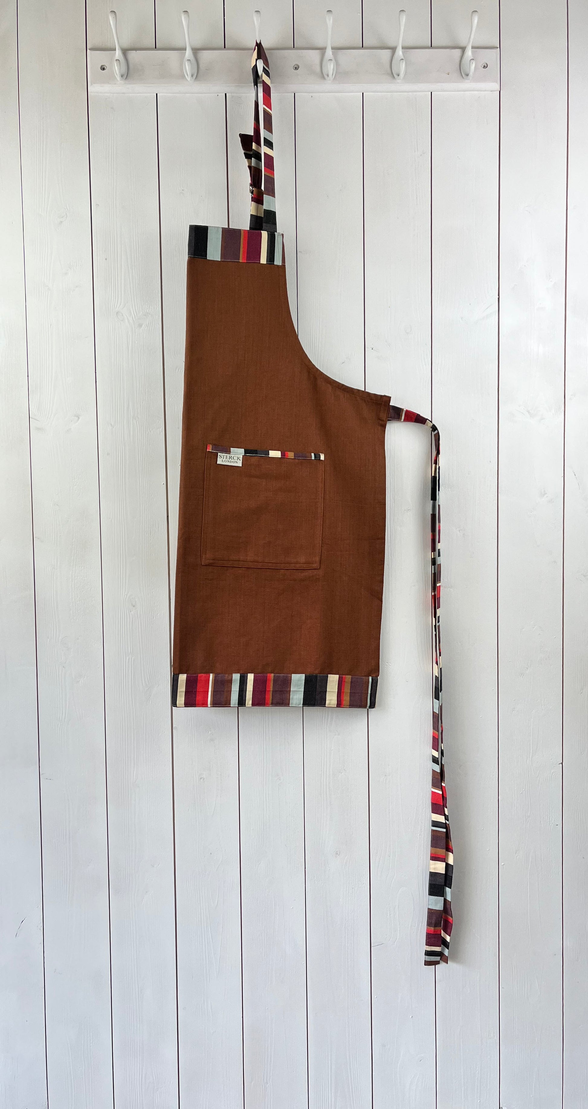 A brown cotton apron with stripy detailing, double front pockets, striped ties and adjustable neck strap. Sterck & Co.