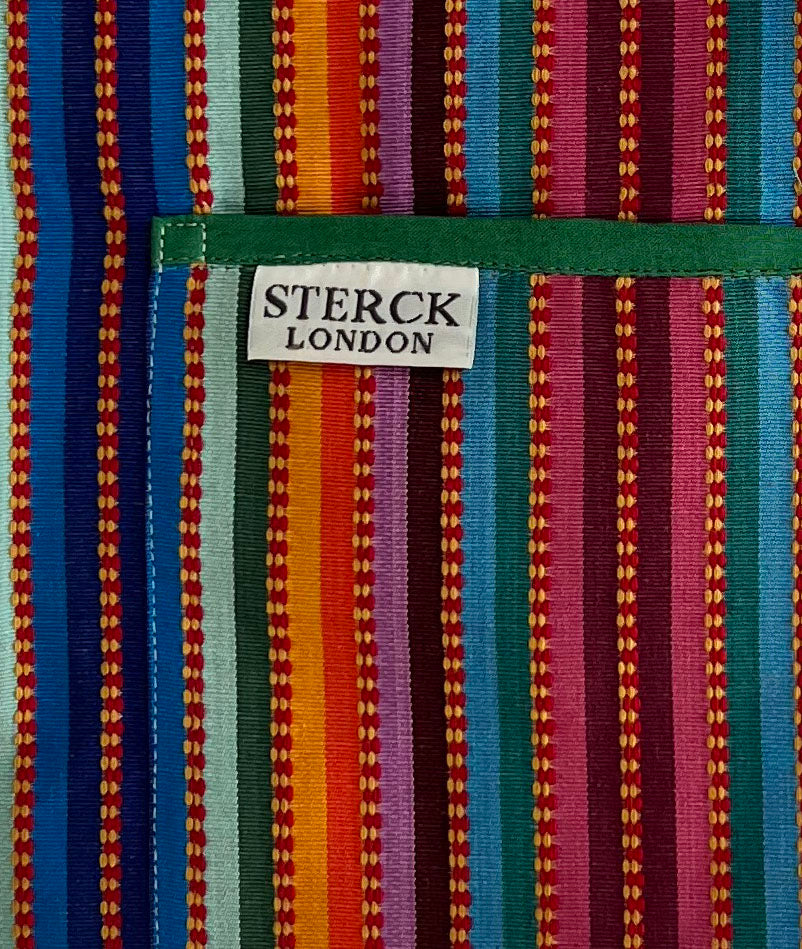 A stripy cotton apron with prominent ticking, two front pockets and adjustable green straps. Close up of fabric and pocket detailing.